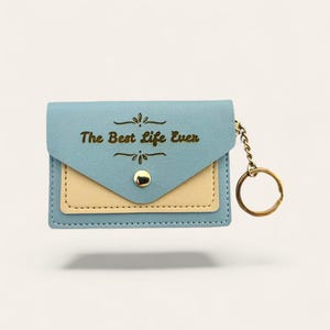 Puede incluir: Cartera azul claro con bolsillo beige y herrajes dorados, que incluyen un cierre a presi&oacute;n y un llavero. El texto "The Best Life Ever" est&aacute; impreso en dorado en la parte delantera.