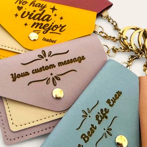 Puede incluir: Primer plano de varios llaveros peque&ntilde;os y coloridos. Los llaveros son de varios colores, incluyendo amarillo, lavanda y azul. Cada llavero tiene un cierre de bot&oacute;n a presi&oacute;n dorado y una cadena dorada. Algunos llaveros tienen el texto "No hay vida mejor", "Your custom message" y "Best Life Ever".