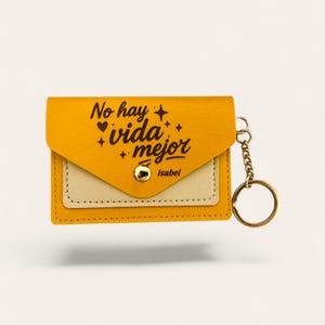 Puede incluir: Un tarjetero de cuero sint&eacute;tico amarillo y beige con un llavero dorado. La cartera tiene un cierre de solapa con un broche dorado. El texto "No hay vida mejor" est&aacute; impreso en la parte delantera, junto con el nombre "Isabel".
