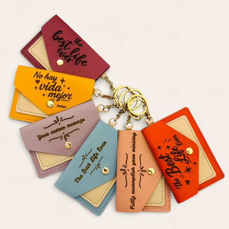Puede incluir: Una colecci&oacute;n de llaveros coloridos con anillos de metal dorado adjuntos. Cada llavero presenta una peque&ntilde;a bolsa rectangular en varios colores, incluyendo borgo&ntilde;a, amarillo y naranja. Las bolsas tienen texto como "The Best Life Ever" y "No hay vida + mejor".