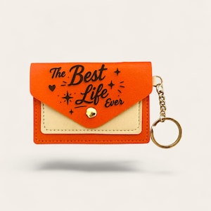 Puede incluir: Monedero llavero naranja y crema con el texto "The Best Life Ever" en escritura negra. El monedero tiene un cierre de bot&oacute;n dorado y una cadena dorada con un llavero. De forma rectangular.