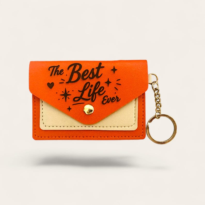 Puede incluir: Monedero llavero naranja y crema con el texto "The Best Life Ever" en escritura negra. El monedero tiene un cierre de bot&oacute;n dorado y una cadena dorada con un llavero. De forma rectangular.
