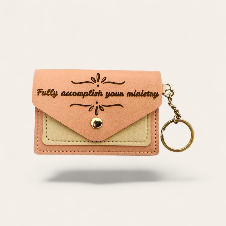 Puede incluir: Un monedero llavero de color melocot&oacute;n con una cadena y un anillo dorados. La cartera tiene una solapa con el texto "Fully accomplish your ministry" y un dise&ntilde;o decorativo. La cartera tiene un bolsillo beige y un cierre de bot&oacute;n a presi&oacute;n dorado.