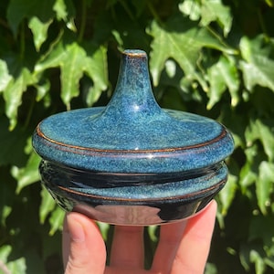 May include: A small, teal blue ceramic jar with a lid. The jar has a rounded, dome-shaped lid with a small, pointed top.