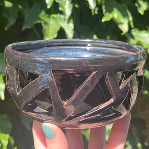 May include: A handmade black ceramic bowl with a geometric cut-out design. The bowl has a dark blue interior and is held against a backdrop of green foliage. The bowl's exterior has a glossy finish.