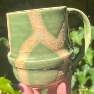 May include: A ceramic mug featuring a glossy sage green glaze with exposed, textured tan clay lines forming a branching pattern. The mug has a distinct horizontal ridge around the middle and a curved handle on the right side, showcasing a handmade aesthetic.