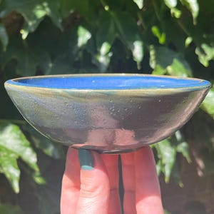May include: A handmade ceramic bowl with a dark exterior and a vibrant blue interior. The bowl is held in front of a backdrop of green foliage. The bowl's rim has a subtle gold accent, adding a touch of elegance to the piece.