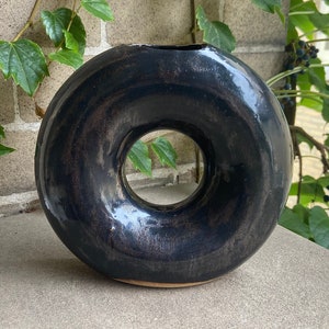 May include: A black ceramic donut-shaped vase with a glossy finish. The vase has a large hole in the center.