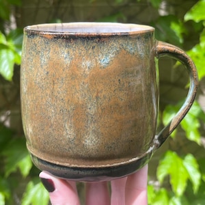 May include: A handmade ceramic mug with a brown and blue glaze. The mug has a rounded body, a curved handle, and a dark base. The mug is held in front of a background of green leaves.
