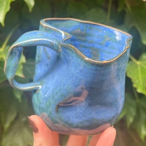 May include: A handmade ceramic mug with a blue glaze and a bird design. The mug has a unique, wavy rim and a curved handle. The mug is held in front of a background of green leaves.