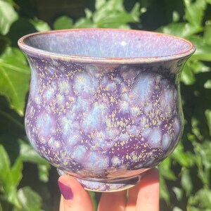 May include: A small ceramic bowl with a rounded silhouette and a flared rim. The exterior features a crystalline glaze with a blend of deep purple, lavender, and light blue tones, accented by small, speckled golden-brown crystalline formations.