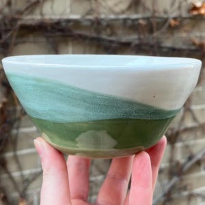 May include: A white ceramic bowl with a green and blue glaze. The bowl is hand-painted with a landscape design.
