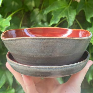 May include: A handmade ceramic bowl and saucer set. The bowl has a square shape with rounded corners and a deep red interior. The exterior is a muted gray-brown color. The saucer is a matching gray-brown. The set is held in front of a green leafy background.