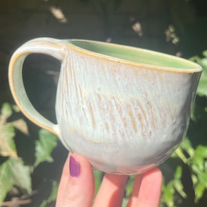 May include: A handmade ceramic mug with a rounded shape and a single handle. The exterior features a textured, light blue and white glaze with visible brown streaks, while the interior is finished in a smooth, pale green glaze with a brown rim.