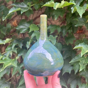 May include: A small, hand-thrown ceramic vase with a rounded body and a narrow neck. The vase is glazed in shades of green and blue, with a subtle white swirl pattern.