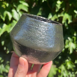 May include: A dark green ceramic cup with a textured, iridescent glaze. The cup has a rounded, slightly flared shape.