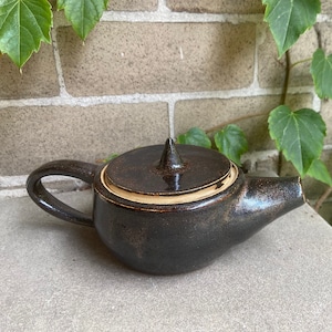 May include: A handmade ceramic teapot with a dark brown glaze. The teapot has a rounded body, a curved handle, and a pointed lid. The rim of the lid is a lighter tan color. The teapot is sitting on a stone surface.