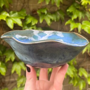 May include: A handmade ceramic bowl with a wavy rim. The bowl features a blend of blue, teal, and dark gray hues, with a glossy finish. The interior has a lighter blue and white pattern. The bowl is held against a backdrop of green leaves.