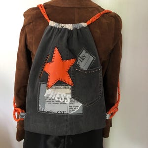 May include: A grey denim drawstring backpack with an orange star applique. The bag has a denim pocket, orange rope straps, and patches with text. The word "PRESS" is visible on one of the patches.