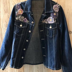 Puede incluir: Chaqueta vaquera azul oscuro con bordados florales en tonos morados, rosas y blancos. La chaqueta tiene botones plateados, dos bolsillos en el pecho y detalles de costuras blancas. Una prenda de abrigo elegante.