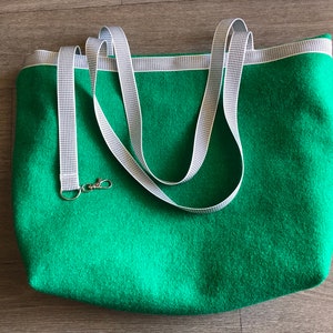 Könnte beinhalten: Eine grüne Filz-Tragetasche mit weißen und grauen, gestreiften Tragegurten. Die Tasche hat einen silbernen Verschluss.