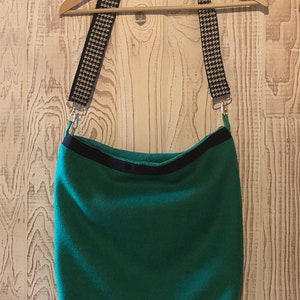 May include: A green shoulder bag with a black trim and a houndstooth patterned strap. The bag is suspended from a wooden hanger against a wood-panelled background. The strap is black and white.