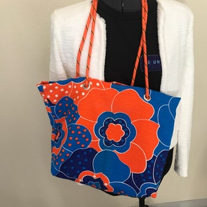 May include: A large orange and blue floral tote bag with a rope handle. The bag is on a white mannequin wearing a white jacket and a black shirt with the text "E UN" on it.