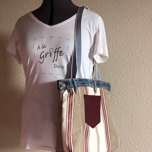 Op de afbeelding: Een gestreepte tote bag met een denim bovenkant en een bordeauxrode leren zak. De tas heeft een gestreept patroon in beige, wit en rood. De tas hangt aan een wit t-shirt met de tekst "À la griffe Paris".