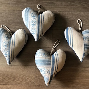 Pode incluir: Quatro almofadas decorativas em forma de coração com um design de tecido listrado e estampado creme e azul. Cada almofada tem uma alça para pendurar. As almofadas estão dispostas sobre uma superfície de madeira.