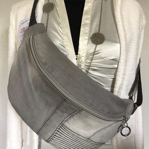 Op de afbeelding: Een grijze denim heuptas met een ritssluiting en een bruine riem. De tas heeft een rechthoekige vorm met stikseldetails en een zilverkleurige metalen ring bevestigd aan de ritssluiting.