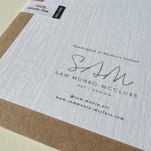 May include: A close-up of a textured white paper with the text "100% plastic free" and "PEEL HERE" on a white label. The paper has the signature "SAM" and the text "SAM MUNRO-MCCLURE ART DESIGN" and website address.