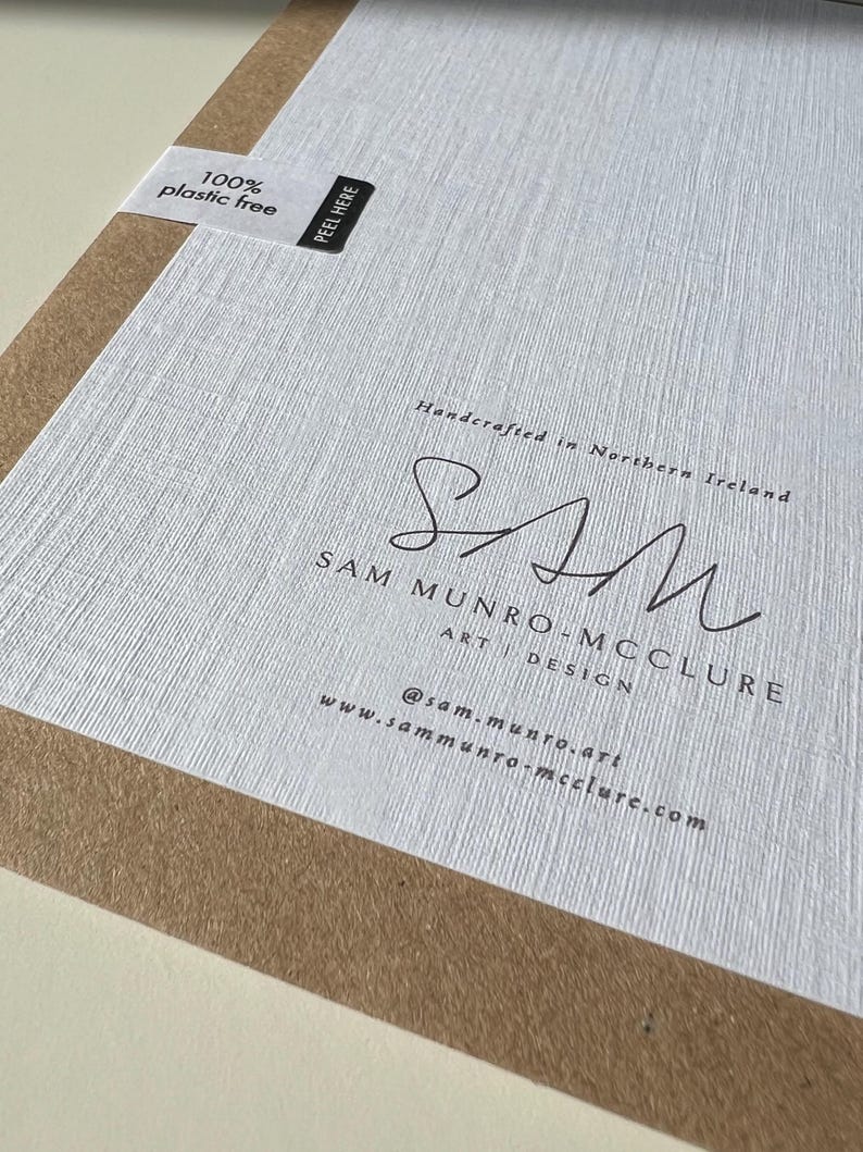 May include: A close-up of a textured white paper with the text "100% plastic free" and "PEEL HERE" on a white label. The paper has the signature "SAM" and the text "SAM MUNRO-MCCLURE ART DESIGN" and website address.
