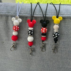 May include: Four beaded lanyards with cartoon mouse head toppers in white, red, black, and yellow. Each lanyard features a variety of beads in coordinating colors, including polka dots and solid colors. Silver-tone lobster clasps are attached.
