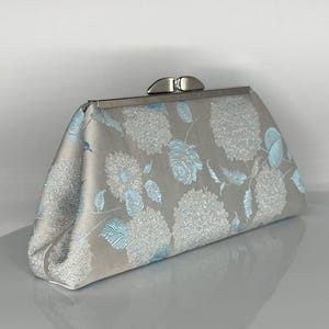 Puede incluir: Un bolso de mano plateado con un estampado floral en azul claro. El bolso rectangular tiene un cierre plateado y un aspecto suave y texturizado. El diseño floral presenta grandes flores y hojas estilizadas.