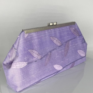 May include: A lavender clutch with a silver clasp. The fabric is embroidered with leaf designs in varying shades of purple. The clutch has a rectangular shape and a structured design.