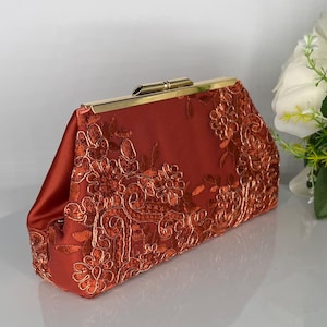 Puede incluir: Un clutch de satén color óxido con intrincados detalles de encaje floral. El clutch presenta un cierre dorado y un diseño estructurado. El bordado de encaje añade un toque de elegancia a este accesorio.