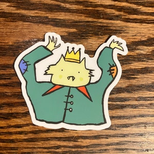 May include: A sticker featuring a cartoon cat wearing a crown and a green jacket with orange and blue patches. The cat has a yellow face, small fangs, and is raising its arms. The sticker has a white border and is on a wooden surface.