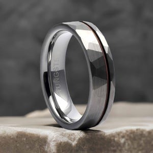 May include: A tungsten carbide ring with a hammered finish and a rose gold inlay. The ring is engraved with the word "TUNGSTEN".