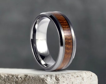 Op de afbeelding: Een zilveren ring van wolframcarbide met een bruine houten inlay. De ring heeft een afgeschuinde rand en een gepolijste afwerking.