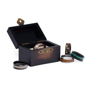 May include: A wooden ring box with a brass latch, open to reveal a silver and copper ring. Five additional rings are shown on a white surface, including a black ring with a silver inlay, a copper ring with a wood inlay, a black ring with a green inlay, and a black ring with a brown inlay.