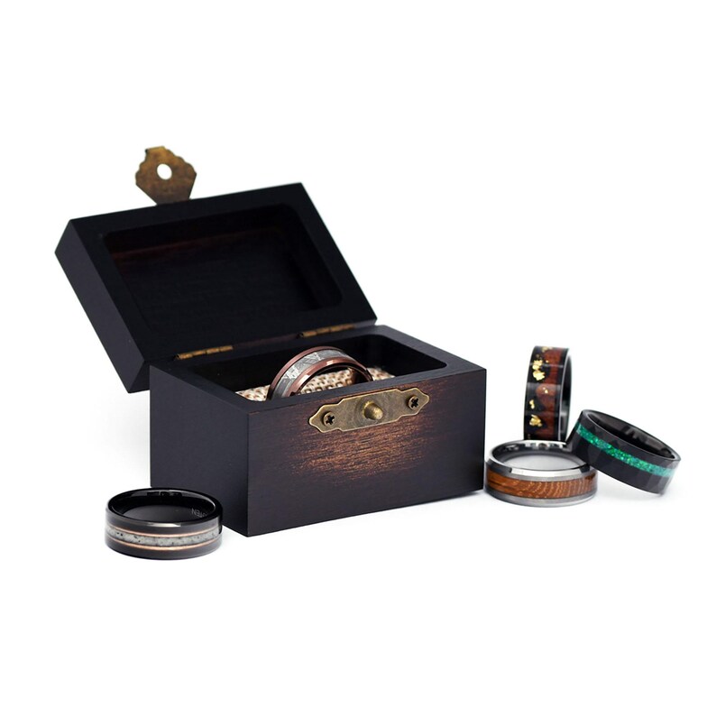 May include: A wooden ring box with a brass latch, open to reveal a silver and copper ring. Five additional rings are shown on a white surface, including a black ring with a silver inlay, a copper ring with a wood inlay, a black ring with a green inlay, and a black ring with a brown inlay.