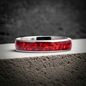 May include: A silver ring featuring a striking red inlay. The band is polished, and the inlay is made up of small, irregular pieces, giving it a textured look. The ring is set against a neutral, textured backdrop.