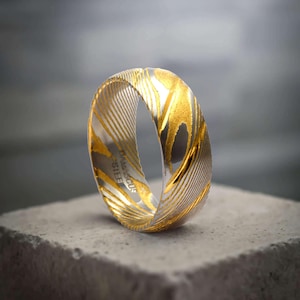 May include: A close-up of a Damascus steel ring with a gold and silver design. The ring has a unique pattern of gold swirls and lines against a silver background. The ring is sitting on a gray surface.