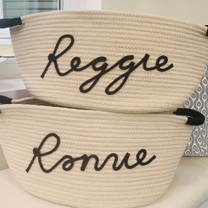 K&ouml;nnte beinhalten: Zwei beigefarbene Korbwaren aus Seil mit schwarzen Griffen und schwarzen Seilbeschriftungen. Der obere Korb sagt "Reggae" und der untere Korb sagt "Ronnie".