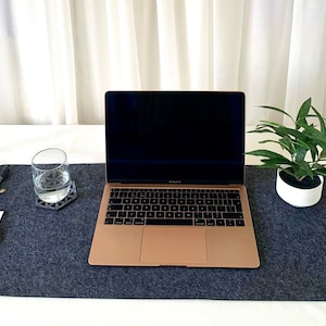 May include: A grey felt desk mat with a laptop computer, a glass of water, a small potted plant, and two pens. The desk mat is on a white surface.