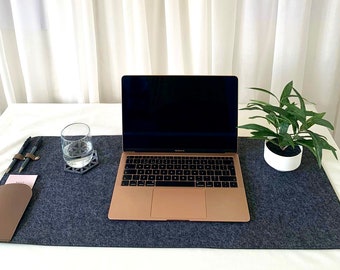 Könnte beinhalten: Eine graue Filzunterlage für den Schreibtisch mit einem Laptop, einem Glas Wasser, einer kleinen Topfpflanze und zwei Stiften. Die Unterlage liegt auf einer weißen Oberfläche.