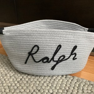 K&ouml;nnte beinhalten: Grauer Korb aus Seil mit schwarzen Seilgriffen und dem Namen "Ralph" auf der Vorderseite gestickt.