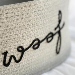 K&ouml;nnte beinhalten: Ein grauer gewebter Aufbewahrungskorb mit dem Wort "woof" in schwarzem Garn gestickt.