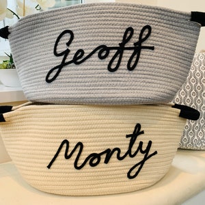 K&ouml;nnte beinhalten: Zwei Aufbewahrungsk&ouml;rbe aus Seil mit schwarzen Griffen. Der obere Korb ist grau mit dem Namen "Geoff", der in Schwarz gestickt ist. Der untere Korb ist cremefarben mit dem Namen "Monty", der in Schwarz gestickt ist.