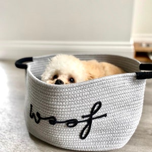 K&ouml;nnte beinhalten: Ein wei&szlig;er Hund ruht in einem grauen Korb aus Seil mit dem Wort "woof" bestickt an der Seite.